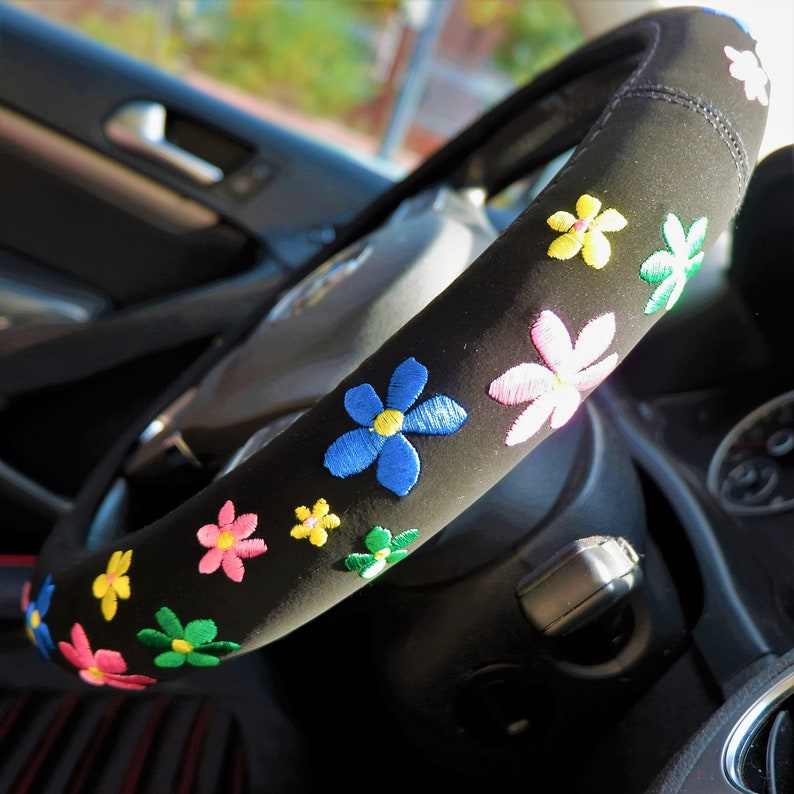 May include: Black steering wheel cover adorned with embroidered flowers in shades of blue, pink, yellow, and green. This cover is designed to fit a standard steering wheel, adding a decorative element to a car's interior.