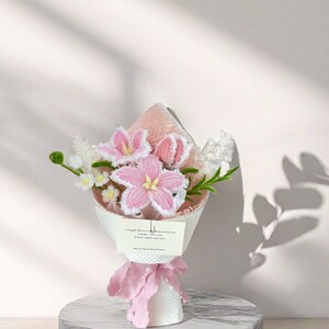 May include: A bouquet of handmade pink and white flowers, wrapped in white paper and tied with a pink ribbon. The bouquet includes lilies and other floral elements. A small card with text is attached to the bouquet.