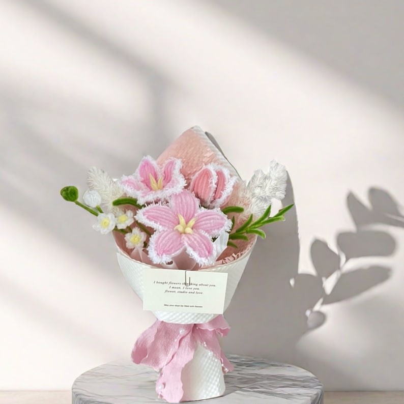 May include: A bouquet of handmade pink and white flowers, wrapped in white paper and tied with a pink ribbon. The bouquet includes lilies and other floral elements. A small card with text is attached to the bouquet.