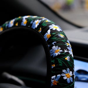 May include: A black steering wheel cover featuring an embroidered pattern of light blue daisy flowers with yellow centers and green stems. The fabric is gathered and fitted around the circular frame of the steering wheel in a vehicle interior.