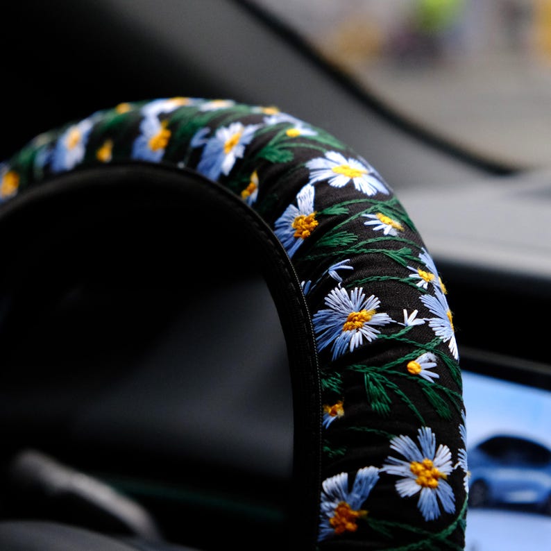 May include: A black steering wheel cover featuring an embroidered pattern of light blue daisy flowers with yellow centers and green stems. The fabric is gathered and fitted around the circular frame of the steering wheel in a vehicle interior.