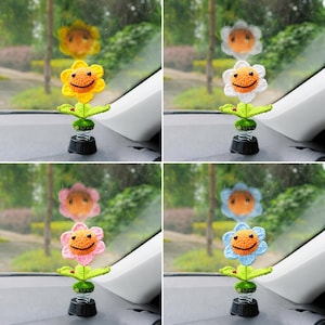 May include: Four car dashboard ornaments. Each features a crocheted flower in yellow, white, pink, or blue, with a smiling face and green leaves. They are mounted on a black base with a spring.