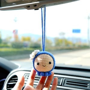 May include: A crocheted doll with a blue hooded head and a white flower on top. The doll has a smiling face with black eyes and a pink mouth. The doll is hanging from a rearview mirror in a car.