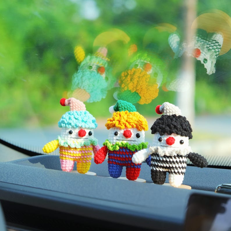 May include: Three handmade crochet clown dolls with colorful outfits and red noses. The clowns have different colored hair and hats, including blue, yellow, and black. They are standing on a gray surface with a blurred green background.