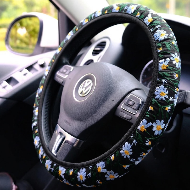 May include: A black steering wheel cover featuring an embroidered pattern of light blue daisy flowers with yellow centers and green stems. The cover is fitted onto a black steering wheel with a silver Volkswagen logo centered on the horn button.