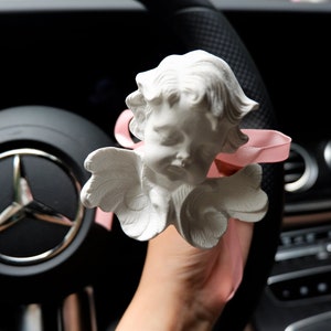 May include: A white ceramic angel figurine with wings and a pink ribbon hanging from the car's rearview mirror.