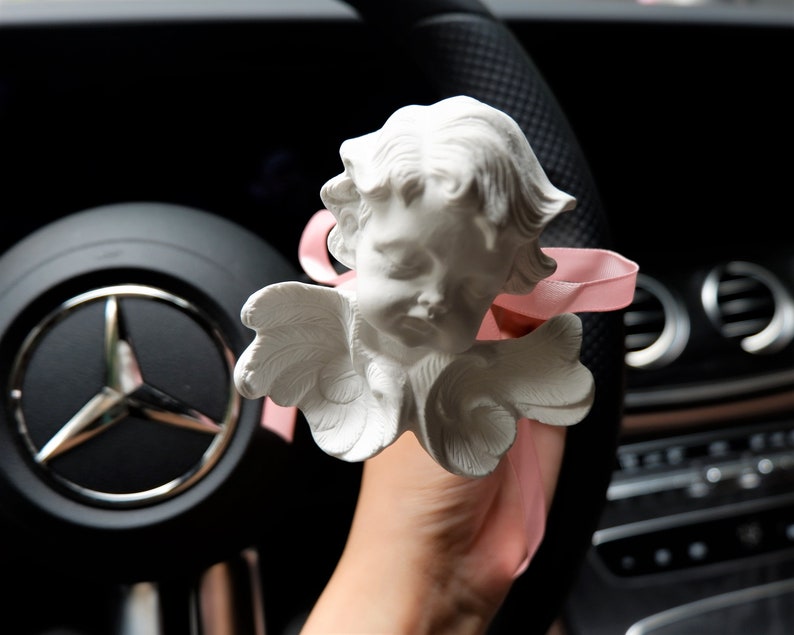 May include: A white ceramic angel figurine with wings and a pink ribbon hanging from the car's rearview mirror.