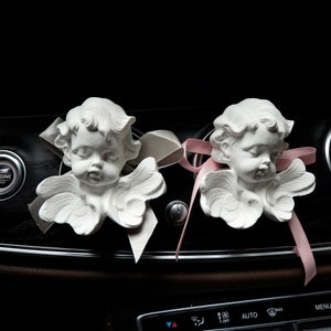 May include: Two white ceramic angel figurines with wings, placed on the dashboard of a car. The angels have closed eyes and are facing forward. The dashboard has a pink ribbon tied around it.