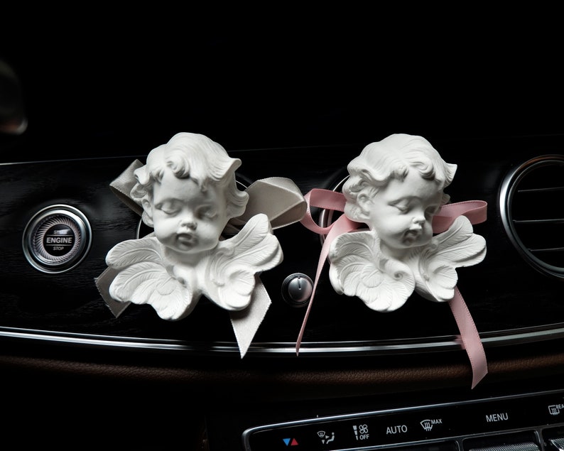 May include: Two white ceramic angel figurines with wings, placed on the dashboard of a car. The angels have closed eyes and are facing forward. The dashboard has a pink ribbon tied around it.