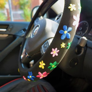 May include: Black steering wheel cover decorated with embroidered flowers in blue, pink, yellow, and green. The cover is designed to fit a standard car steering wheel.