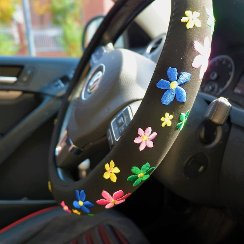 May include: Black steering wheel cover decorated with embroidered flowers in blue, pink, yellow, and green. The cover is designed to fit a standard car steering wheel.