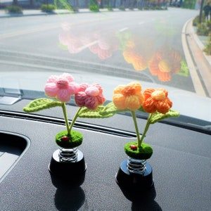 May include: Two car dashboard decorations featuring crocheted flowers on spring-loaded bases. The flowers are pink and orange.