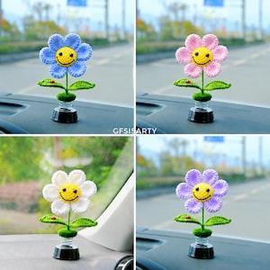 May include: Four crochet flower car decorations in blue, pink, white, and purple. Each flower has a yellow smiley face centre, green leaves, and a black spring base. The text "GFISISARTY" is visible.