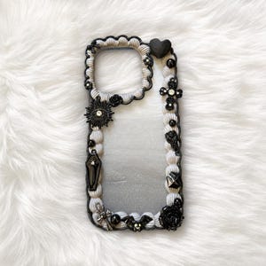 May include: A decorative phone case with a black and white gothic design. The case features a black heart, crosses, roses, and a coffin. The border is decorated with black and white embellishments and rhinestones.