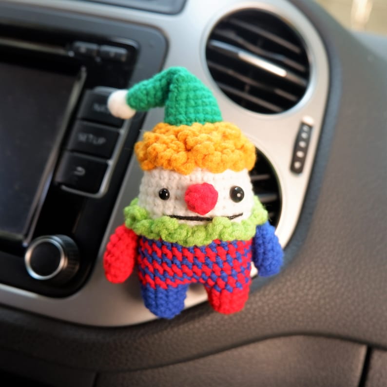 May include: A crocheted clown doll with a green hat, yellow hair, white face, red nose, and a green, red, and blue patterned body. The doll is hanging from the air vent of a car.