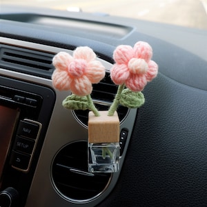Puede incluir: Un ambientador de coche con dos flores de crochet rosas y hojas verdes en una botella de vidrio transparente con tapa de madera.