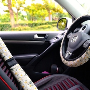May include: A car steering wheel cover and a seat belt shoulder pad cover, both featuring a cream-colored fabric with a repeating pattern of white daisy flowers, yellow centers, and green stems, installed inside a vehicle with a black interior.