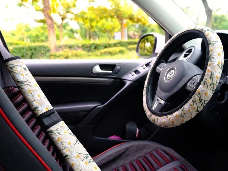 May include: A car steering wheel cover and a seat belt shoulder pad cover, both featuring a cream-colored fabric with a repeating pattern of white daisy flowers, yellow centers, and green stems, installed inside a vehicle with a black interior.
