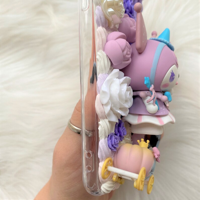 Puede incluir: Una funda transparente para tel&eacute;fono decorada con un personaje 3D morado y blanco, flores y un carruaje de calabaza rosa. El personaje lleva un sombrero azul y un vestido blanco. La funda est&aacute; decorada con flores blancas, moradas y rosas.