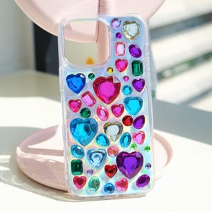May include: Clear phone case adorned with an array of colorful, jewel-like embellishments. The design features heart-shaped and rectangular gems in shades of blue, pink, green, and purple, creating a sparkling, eye-catching accessory.