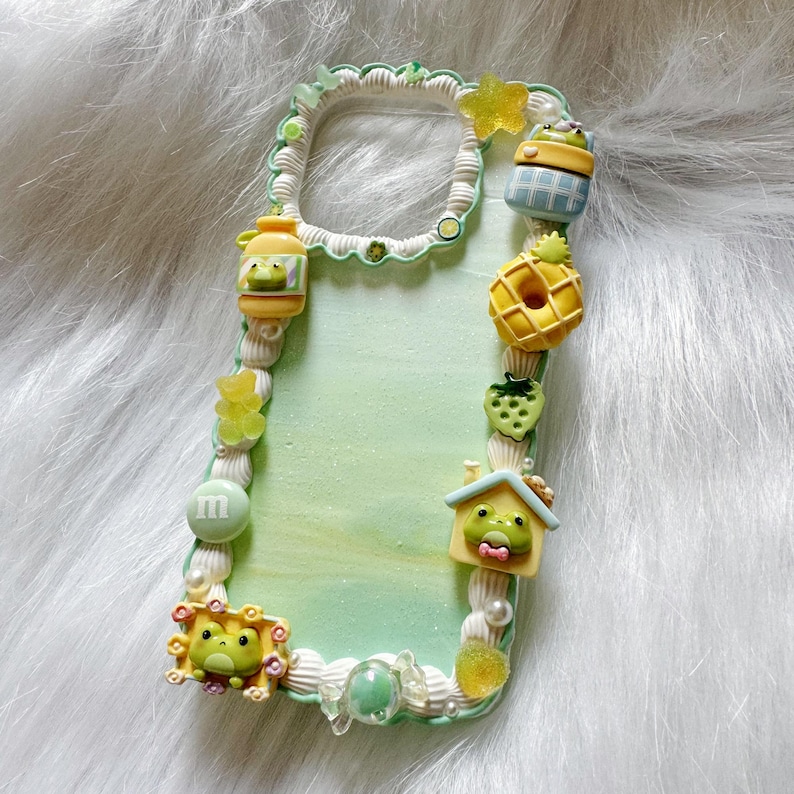 May include: A decorative phone case with a light green background and a white, textured border. The case is adorned with various colorful, miniature embellishments, including a frog, a pineapple, a strawberry, and a house.