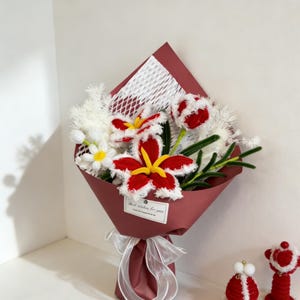 May include: A bouquet of handmade flowers wrapped in burgundy paper, tied with a white ribbon. The bouquet features red and white crocheted lilies and tulips, with green leaves and white filler. A small tag reads "Best wishes for you."
