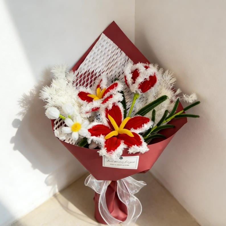 May include: A bouquet of handmade flowers, featuring red and white lilies, tulips, and daisies. The flowers are wrapped in burgundy paper and tied with a white ribbon. The bouquet includes the text "Best wishes for you."
