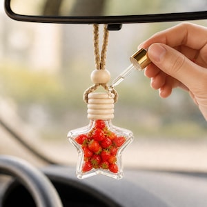 Pode incluir: Um frasco de vidro para ambientador de carro em forma de estrela, preenchido com pequenos berloques vermelhos em forma de morango. O frasco possui uma tampa de madeira clara e um cordão trançado. Um conta-gotas dourado está posicionado no topo.