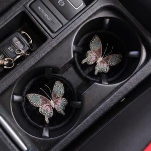 May include: Two black car coasters with embroidered butterfly designs. The butterflies feature green, pink, and purple thread. The coasters are in a car's center console cup holders, next to a car key.