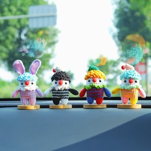 May include: Four colorful, handmade crochet dolls with whimsical designs. One doll has bunny ears, while the others resemble clowns with various hat styles. Each doll stands on a small wooden base, showcasing a variety of vibrant colors.