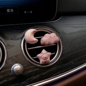 May include: Three wooden car air vent decorations: a crescent moon, a smiling cloud, and a star with a face. The decorations are light brown and pink, and are placed in a car's air vent.