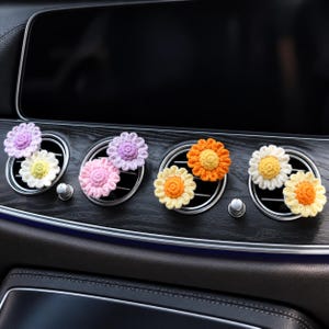 May include: A set of eight small, round, crocheted flower ornaments attached to the circular air vents of a vehicle dashboard. The flowers feature various colour combinations including lavender, white, yellow, pink, and orange petals.