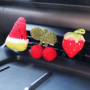 Puede incluir: Ambientadores de coche de frutas hechos a ganchillo. El conjunto incluye una rodaja de sandía, cerezas y una fresa. La sandía es roja, blanca y verde. Las cerezas son rojas con hojas verdes. La fresa es roja con la parte superior verde.