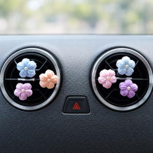 May include: Five small, crocheted flower ornaments in pastel shades of light blue, peach, soft pink, and lavender, each featuring a tiny white bead at the centre. These decorative floral accents are attached to the black circular air vents of a car.