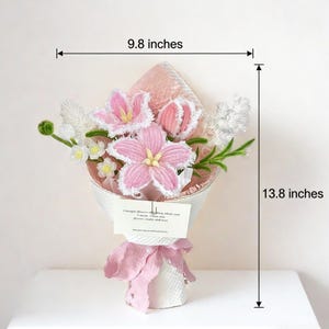 May include: A bouquet of pink and white crocheted flowers, measuring 9.8 inches wide and 13.8 inches tall, wrapped in white paper and tied with a pink ribbon. A small card with text is attached. The flowers include lilies and other blossoms.