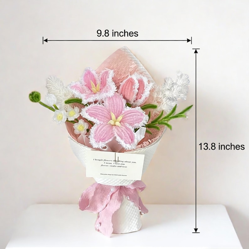 May include: A bouquet of pink and white crocheted flowers, measuring 9.8 inches wide and 13.8 inches tall, wrapped in white paper and tied with a pink ribbon. A small card with text is attached. The flowers include lilies and other blossoms.