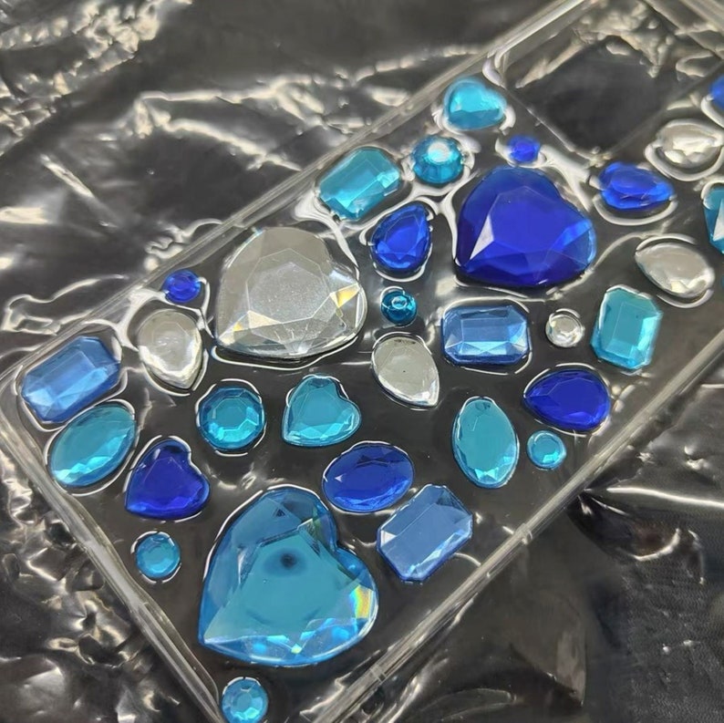 May include: Clear phone case decorated with an assortment of blue and silver-colored rhinestones. The rhinestones are various shapes, including hearts, ovals, and rectangles. The case is on a dark, textured surface.