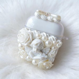 May include: White AirPod case with a decorative cherub, roses, and faux pearls. The case has a clear top and a pearl-like accent. The design is a romantic, ornate style.