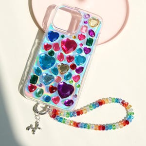 May include: Clear iridescent phone case adorned with colorful heart-shaped and other gemstone embellishments. A rainbow beaded strap is attached to the case with a silver charm. The case features a variety of colors, including red, pink, blue, green, and purple.