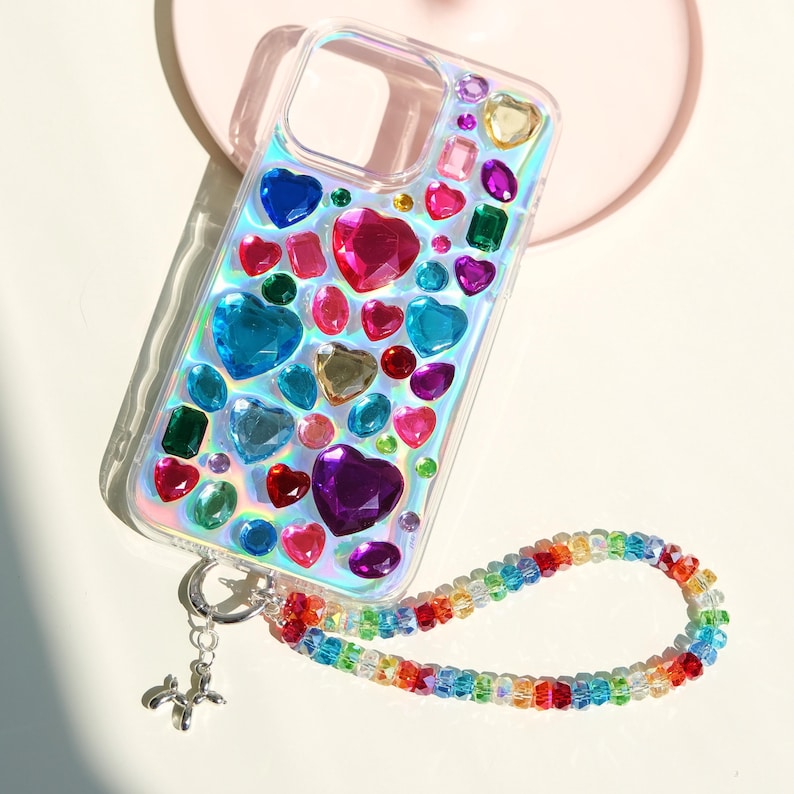 May include: Clear iridescent phone case adorned with colorful heart-shaped and other gemstone embellishments. A rainbow beaded strap is attached to the case with a silver charm. The case features a variety of colors, including red, pink, blue, green, and purple.