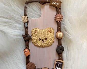 Puede incluir: Funda de teléfono marrón con un centro rosa claro, decorada con varios adornos temáticos de comida. Presenta un osito de peluche, chocolate, gofres y un letrero que dice "Delicious coffee".