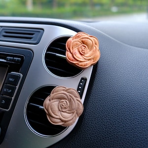 May include: Two wooden rose-shaped car air fresheners. One is a light brown color, and the other is a darker brown. They are attached to the car's air vents, which are silver and black. The dashboard is black.