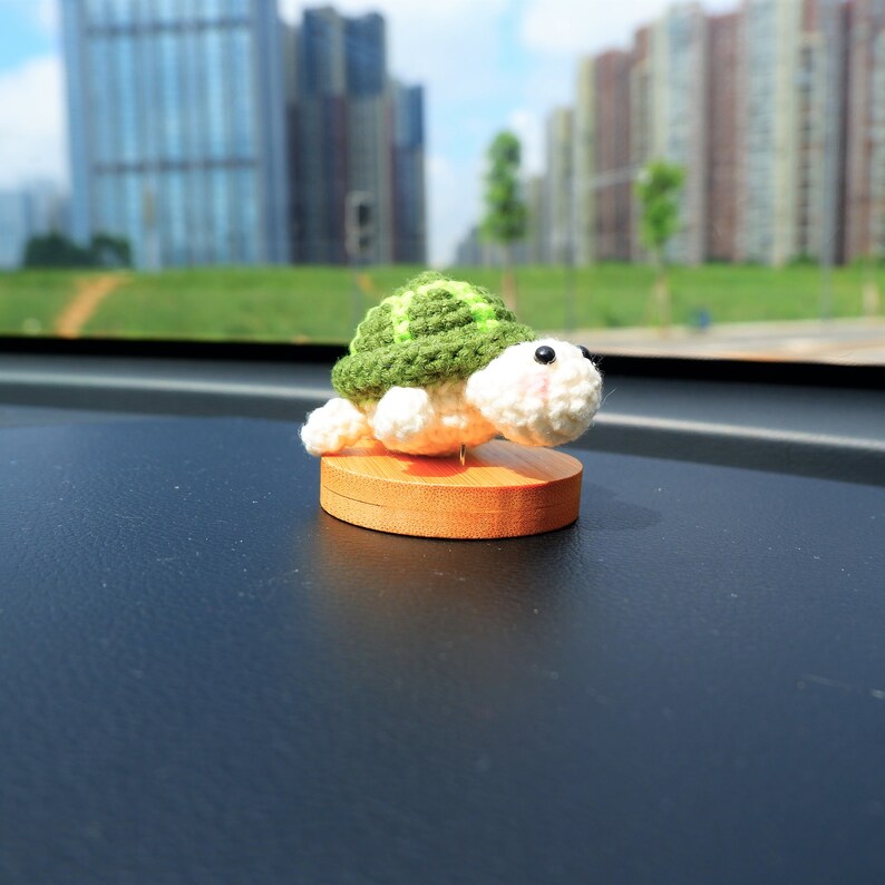 May include: A handcrafted, crocheted turtle figurine with a green shell and white body, mounted on a small, round wooden base. The turtle has black eyes and pink cheeks. The figurine is designed to sit on a dashboard.