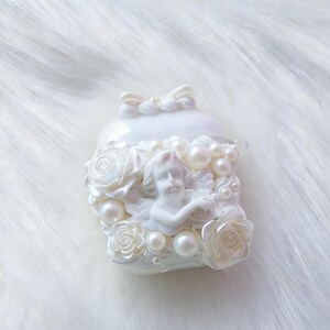 May include: A white, pearlescent trinket box with a sculpted cherub playing a harp, surrounded by roses and faux pearls. The box has a bow on top, creating a romantic and elegant aesthetic.