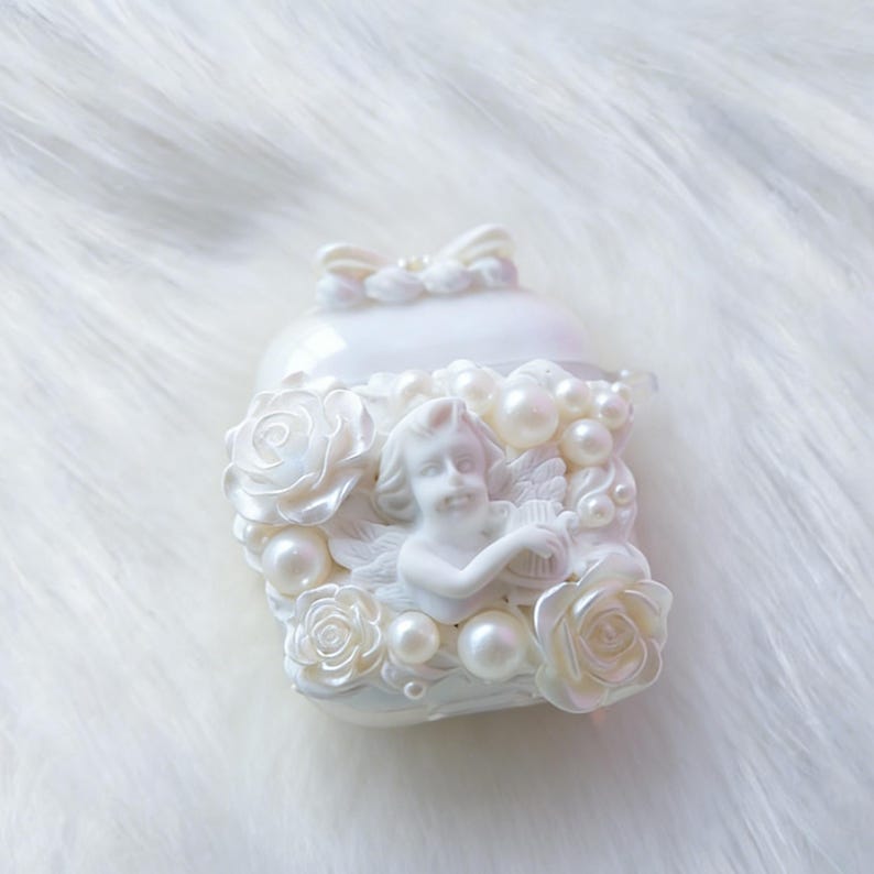 May include: A white, pearlescent trinket box with a sculpted cherub playing a harp, surrounded by roses and faux pearls. The box has a bow on top, creating a romantic and elegant aesthetic.