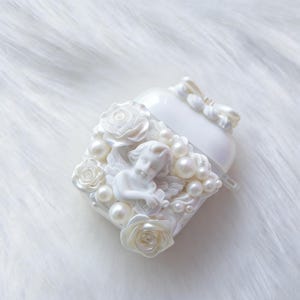 May include: White heart-shaped case with a sculpted cherub, roses, and faux pearls. The case has a bow on top and is set against a white, fluffy background. The case is likely for small electronics.