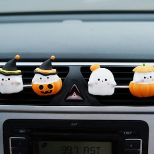 May include: Four Halloween-themed car vent decorations. Two are white ghosts with black witch hats, one is a white ghost with an orange ball, and the last is an orange pumpkin with a green stem. They are attached to a car vent.