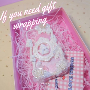 Puede incluir: Una funda de tel&eacute;fono blanca y rosa con un marco decorativo, flores y un dise&ntilde;o de &aacute;ngel. La funda est&aacute; en una caja de regalo rosa con papel de seda rosa. La caja tambi&eacute;n incluye una etiqueta a cuadros rosa y blanca con el texto "LOVE THINGS".