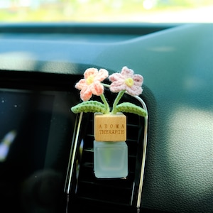 May include: A car air freshener with two crocheted pink flowers, green leaves, and stems. The flowers are in a small wooden block with "AROMA THERAPIE" and a frosted glass bottle. The air freshener is attached to a car vent, a decorative item.
