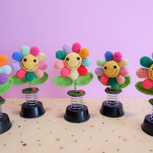 May include: Five colorful crocheted flowers with smiling faces on spring-loaded bases. Each flower has a different color combination of pink, purple, green, yellow, and blue.